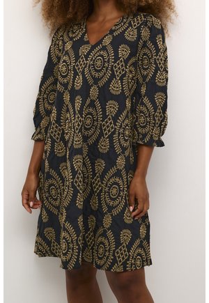 Frau trägt ein schwarzes knielanges Kleid mit goldenen, bestickten Blumen- und runden Mustern, mit dreiviertel Ärmeln und V-Ausschnitt.