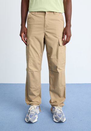 Beige cargo pants made of lightweight fabric with multiple pockets and a relaxed fit, paired with gray and white athletic shoes.
