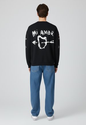 Sweatshirt noir avec graphique blanc "Mi Amor" et design de flèche dans le dos. Associé à un pantalon à motifs bleu clair et des baskets blanches.