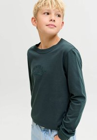 Majica dugih rukava od tamnozelene pamučne tkanine s teksturiranim logotipom izvezenim na prednjoj strani, okruglim izrezom i uskim manžetama. Uparena s svijetloplavim trapericama.