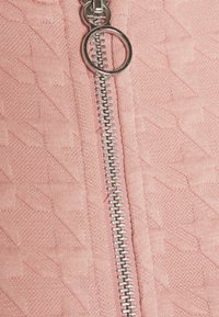 Primo piano di una cerniera metallica argento con un anello rotondo su un tessuto rosa testurizzato con un sottile motivo diagonale.