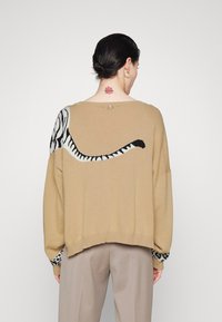 Pull beige avec un motif de queue de tigre en noir et blanc, doté d'une coupe ample et de poignets côtelés, porté sur un pantalon beige clair.