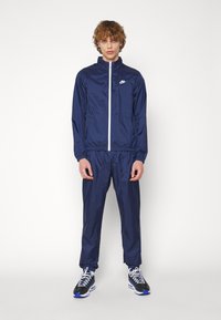 Σκούρο μπλε αθλητική φόρμα της Nike από ελαφρύ ύφασμα; διαθέτει φερμουάρ στη ζακέτα με λευκό φερμουάρ και αντίστοιχες παντελόνα με ελαστική μέση.