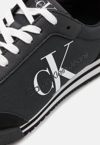 �Černé tenisky s vrchní částí z mesh materiálu, bílé tkaničky a výrazným logem CK. Měkká textura s kontrastními bílými pruhy na gumové podrážce.