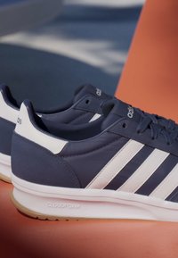Par de zapatillas Adidas azul marino y blancas con tres franjas, medias suelas Cloudfoam, suelas exteriores de goma texturizadas y cierre con cordones sobre una superficie naranja.