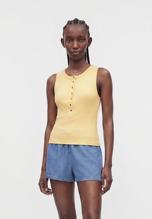 Γυναίκα με πλεξούδες, που φοράει ένα κίτρινο αμάνικο τοπ με κουμπιά και μπλε σορτς με ρίγες, στέκεται μπροστά σε ένα απλό φόντο.