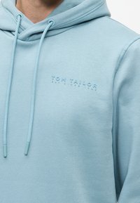 Ανοιχτό μπλε φούτερ από μαλακό ύφασμα, με μπροστινή τσέπη, κορδόνια και ανάγλυφο λογότυπο "TOM TAILOR" στο στήθος.