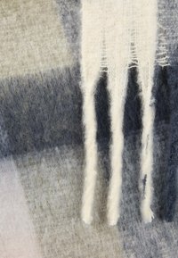 Uld tørklæde med et ternet mønster i grå, blå og cremefarve. Har frynser i kanten med tre fremtrædende garntråde hængende. Blød tekstur.