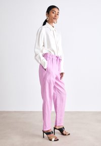 Chemise blanche à manches longues avec col, associée à un pantalon rose clair à taille élastique. Sandales à talons noires et beige ; textures lisses, design minimal.