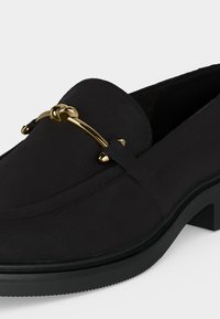 Mocassin en cuir noir avec petit talon et décoration en métal doré en forme de mors de cheval sur la partie supérieure avant.