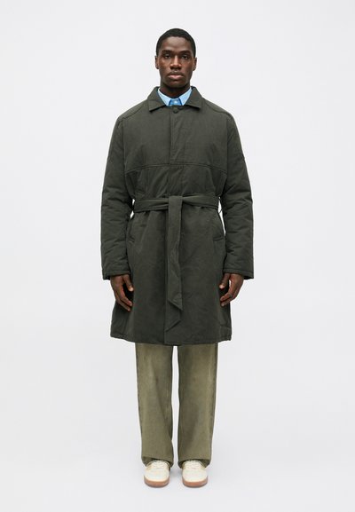 Trench coat lung de culoare verde închis, cu centură și guler, asortat cu pantaloni verzi deschis și adidași albi. Material neted, design structurat, croială casual.