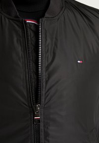 Sort bomberjakke lavet af glat, holdbart stof. Har en frontlynlås og et lille logo-patch med røde, hvide og blå detaljer.