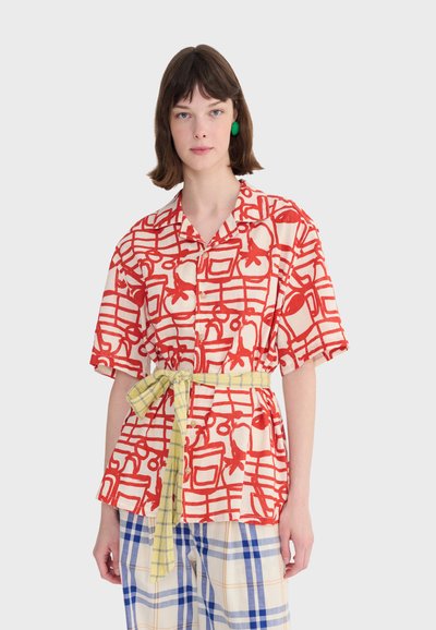 Femme portant une chemise à manches courtes rouge à motifs, une ceinture à carreaux jaune et un pantalon à carreaux bleu et blanc sur un fond uni.