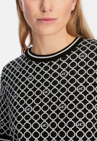 Schwarzer und weißer gemusterter Pullover mit kreisförmigen Designs, ausgestattet mit einem gerippten Rundhalsausschnitt und strukturiertem Strickstoff. Akzente beinhalten Logodetails.