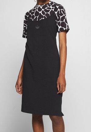 Persoon die een zwarte mouwloze jurk draagt over een zwart-wit giraffenpatroon shirt met korte mouwen, staand tegen een effen achtergrond.