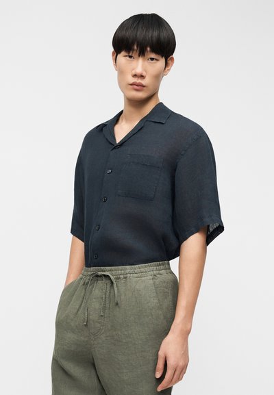 Chemise en lin bleu marine à manches courtes avec col boutonné, poche poitrine unique et coupe décontractée, associée à un pantalon olive à cordon de serrage.
