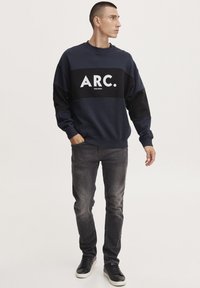 Felpa navy con un blocco orizzontale nero che presenta il logo bianco "ARC.", abbinata a jeans grigi e sneakers nere. Tessuto morbido, vestibilità casual.
