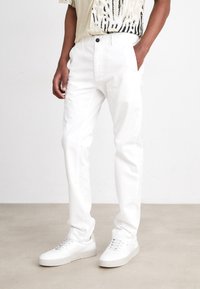 White cotton pants with a straight fit, featuring side pockets and a button closure. Worn with white sneakers on a beige floor.