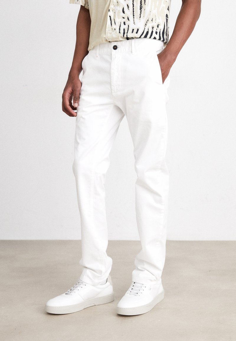 White cotton pants with a straight fit, featuring side pockets and a button closure. Worn with white sneakers on a beige floor.