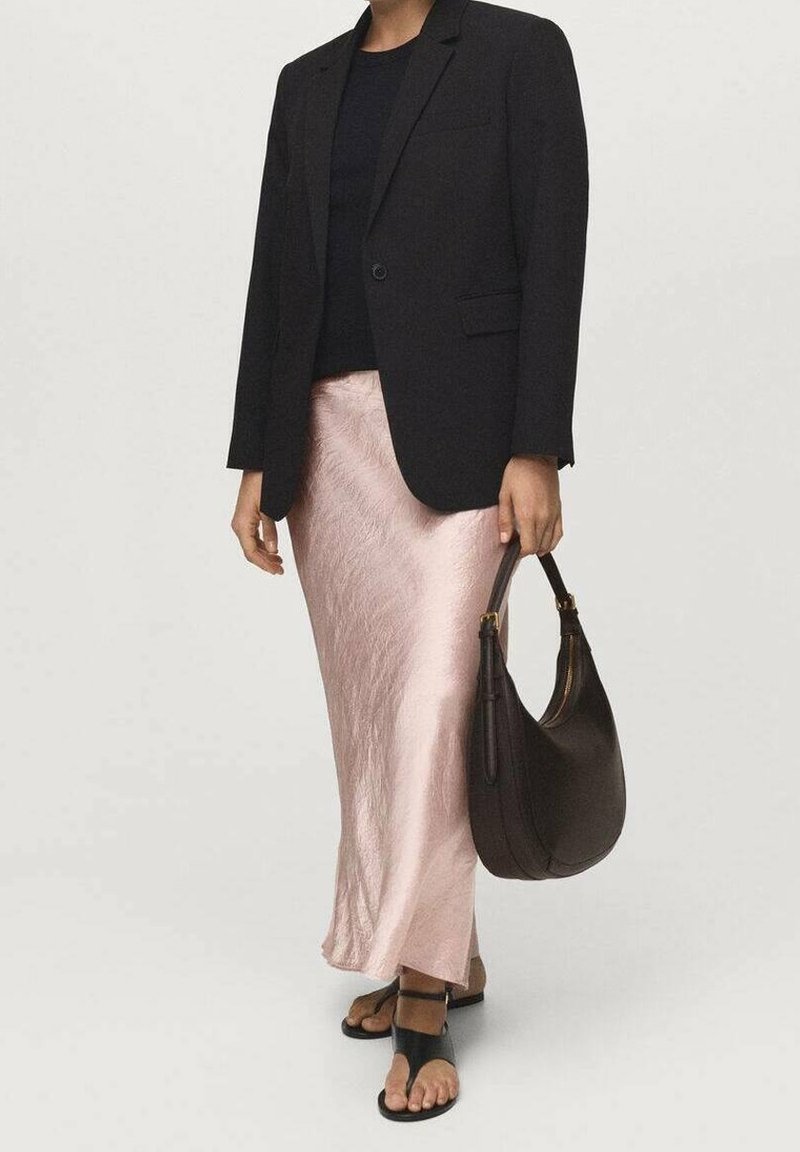 Femme portant un blazer noir, une jupe en satin rose pâle, des sandales noires, et tenant un sac à main marron foncé sur un fond uni.