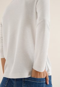 Maglione grigio chiaro con texture, maniche 3/4, vestibilità comoda, orlo curvo e spacchi laterali, abbinato a jeans in denim blu.