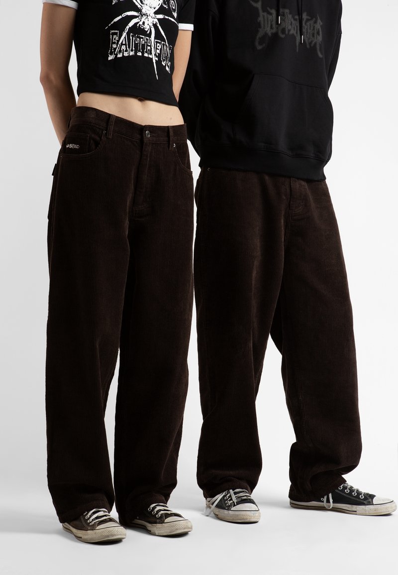 Deux personnes portant des pantalons larges en velours côtelé marron foncé et des baskets noires, l'une avec un t-shirt graphique court et l'autre avec un sweat à capuche noir.