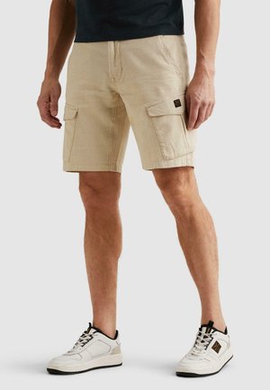 Mann, der beige Cargoshorts und weiße Sneakers trägt, steht vor einem schlichten Hintergrund.