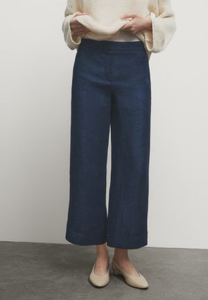 Persoon die brede donkerblauwe broek, crèmekleurige gebreide trui en beige schoenen met lage hak draagt, staande op een grijze vloer tegen een witte muur.