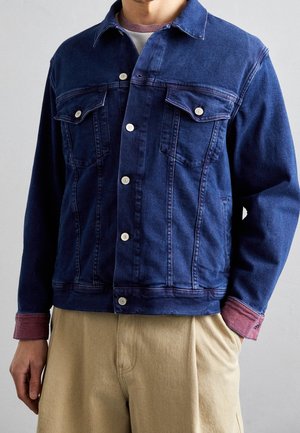 Uomo che indossa una giacca di jeans blu scuro con bottoni frontali, tasche al petto, polsini rossi, pantaloni beige e una camicia a strati bianca e bordeaux.