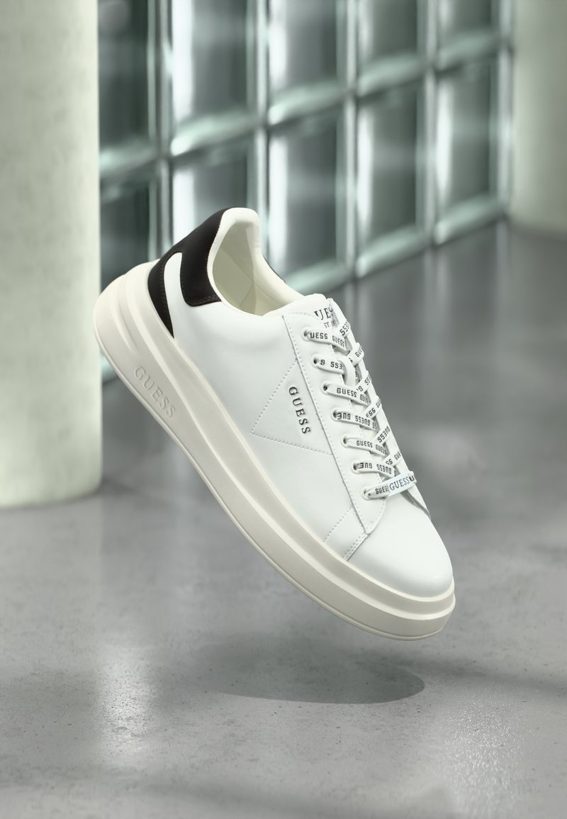 Fehér és fekete GUESS sneaker márkázott fűzőkkel, egy szürke padló fölött lebegve, egy üvegtégla fal közelében és egy henger alakú oszlop mellett.