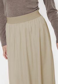 Jupe maxi beige fluide avec une taille élastique, fabriquée en tissu léger. Présente des plis doux pour plus de texture et de mouvement.