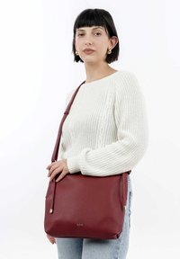 Sac en bandoulière rouge en cuir avec une finition texturée, une sangle réglable et un accent en métal argenté. Porté avec un pull en tricot crème.