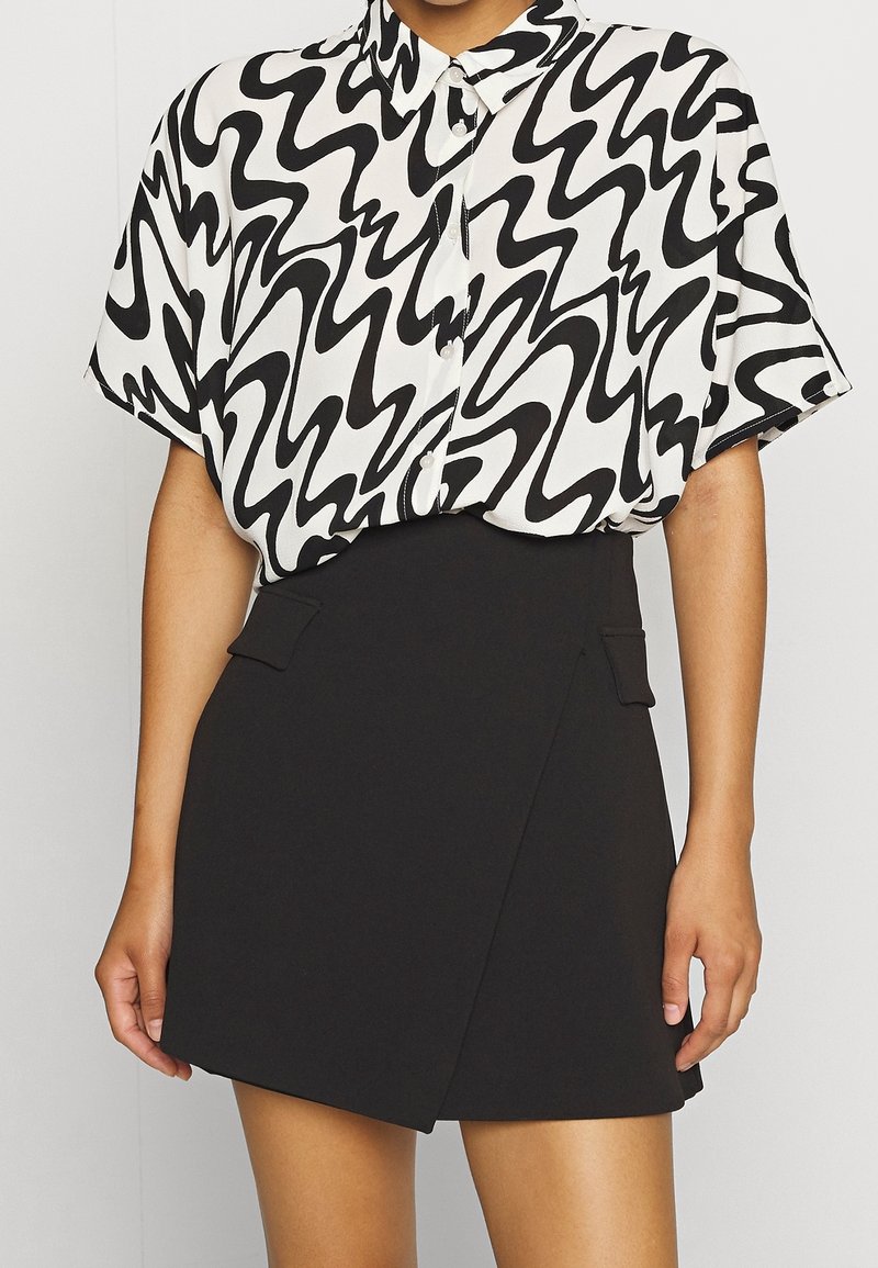 Černobílá košile s abstraktním potiskem, krátkými rukávy a límečkem, doplněná černou zavinovací sukní s bočními kapsami.