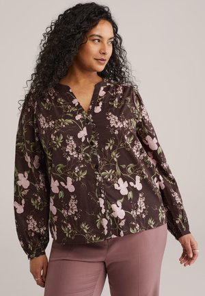 Göndör hajú nő, sötét virágos gombos blúzt és mályva nadrágot visel, oldalra néz egyszerű háttér előtt.