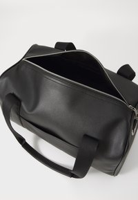 Schwarze Sporttasche mit glatter, matter Oberfläche, Reißverschlussdeckel, zwei Griffen und minimalistischem Design. Das Innere ist ungefüttert und dunkel.