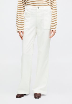 Femme portant un pantalon blanc taille haute à jambes larges avec poches plaquées à l'avant, un haut à manches longues rayé marron, et des chaussures marron à bout carré.