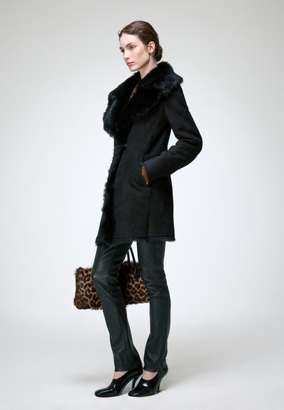 Femme portant un manteau noir à col en fourrure, un pantalon en cuir noir, des talons hauts noirs, et tenant un sac à main imprimé léopard, debout de profil.