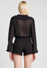 Schwarze transparente Bluse mit Punkten und Rüschenkragen, kombiniert mit schwarzen Shorts mit Gesäßtaschen und figurbetontem Schnitt.