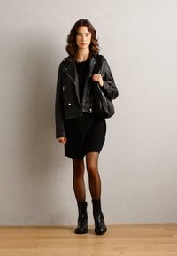 Black leather motorcycle jacket over a black dress, paired with black ankle boots and a slouchy black handbag, standing on a wooden floor.