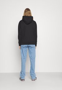 Felpa nera con cappuccio regolabile, abbinata a jeans chiari svolazzanti e sneaker nere con dettagli bianchi. Sfondo semplice.