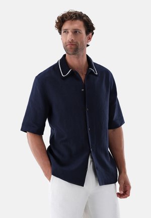 Mann mit lockigem Haar trägt ein marineblaues kurzärmeliges Hemd mit weiß eingefasstem Kragen und weiße, strukturierte Hose, Hände in den Taschen.