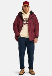 Chaqueta de plumas burdeos con acentos negros, sudadera beige con logo, pantalones cargo azul marino y botas de cordones marrones. Atuendo casual para el exterior.