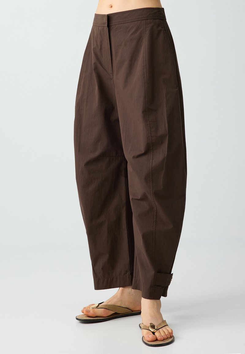 Personne portant un pantalon marron ample avec les chevilles retroussées et des tongs beiges, debout devant un fond clair uni.