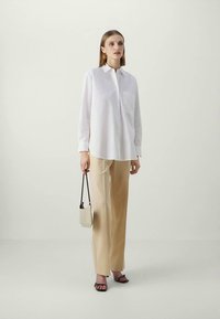 White collared shirt with a chest pocket, paired with beige wide-leg trousers. Model holds a small beige handbag and wears black sandals.