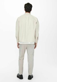 Camicia beige chiaro a maniche lunghe con colletto a bottoni, abbinata a pantaloni grigi a righe e scarpe nere. La camicia presenta polsi con bottoni.