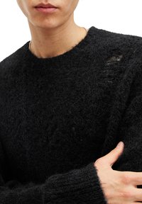Czarny sweter dzianinowy z fakturowanym, puszystym wykończeniem oraz widoczną dziurą na lewym ramieniu. Okrągły dekolt i długie rękawy.