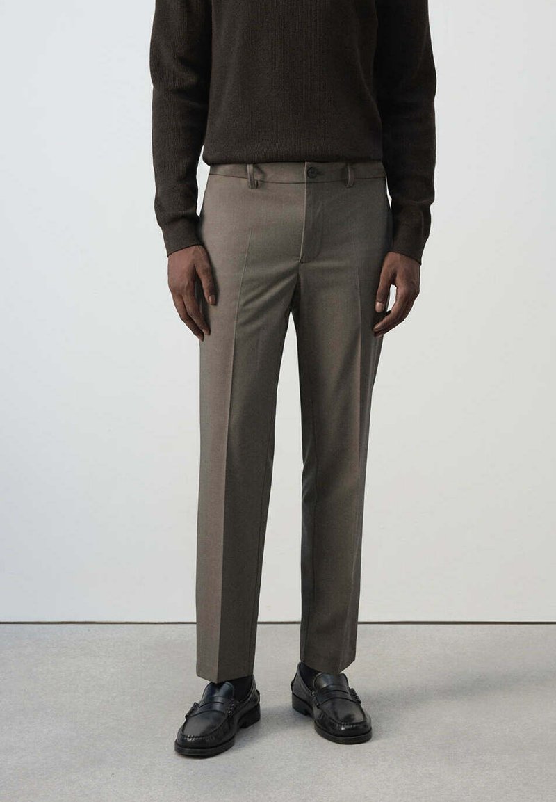 Homme portant un pantalon marron sur mesure, des mocassins noirs et un haut à manches longues marron foncé, debout devant un fond clair uni.