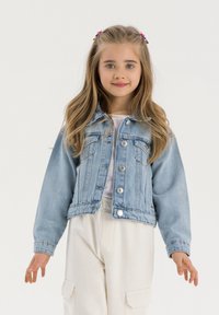 Veste en denim bleu clair avec un design court, fermeture à boutons sur le devant et deux poches poitrine. Portée sur une chemise blanche et un pantalon crème.