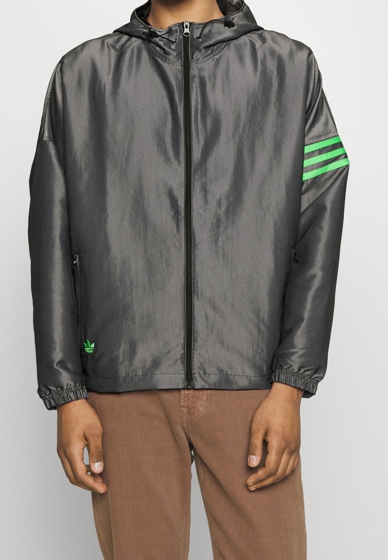 Muž v šedé zip-up bundě se zelenými pruhy na levé paži a odpovídajícím zeleným logem poblíž levé kapsy, ve spojení s hnědými kalhotami.