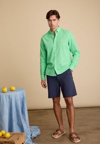 Lichtgroen overhem shirt, marineblauwe shorts en bruine sandalen. Accessoires zijn citroenen op een lichtblauwe tafellaken, eenvoudig ontwerp en texturen.